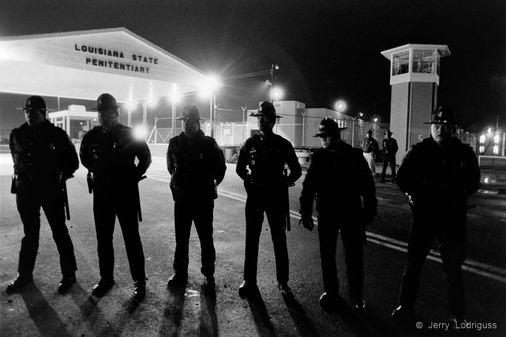 A squad of Louisiana State Troopers stands between the front gate of the State Penitentiary at Angola and a small group of death-penalty protesters keeping a prayer vigil here 12/4/83 as Tobert Wayne Williams was executed by electrocution. The Watchtower at right overlooks the celblock containing death row.