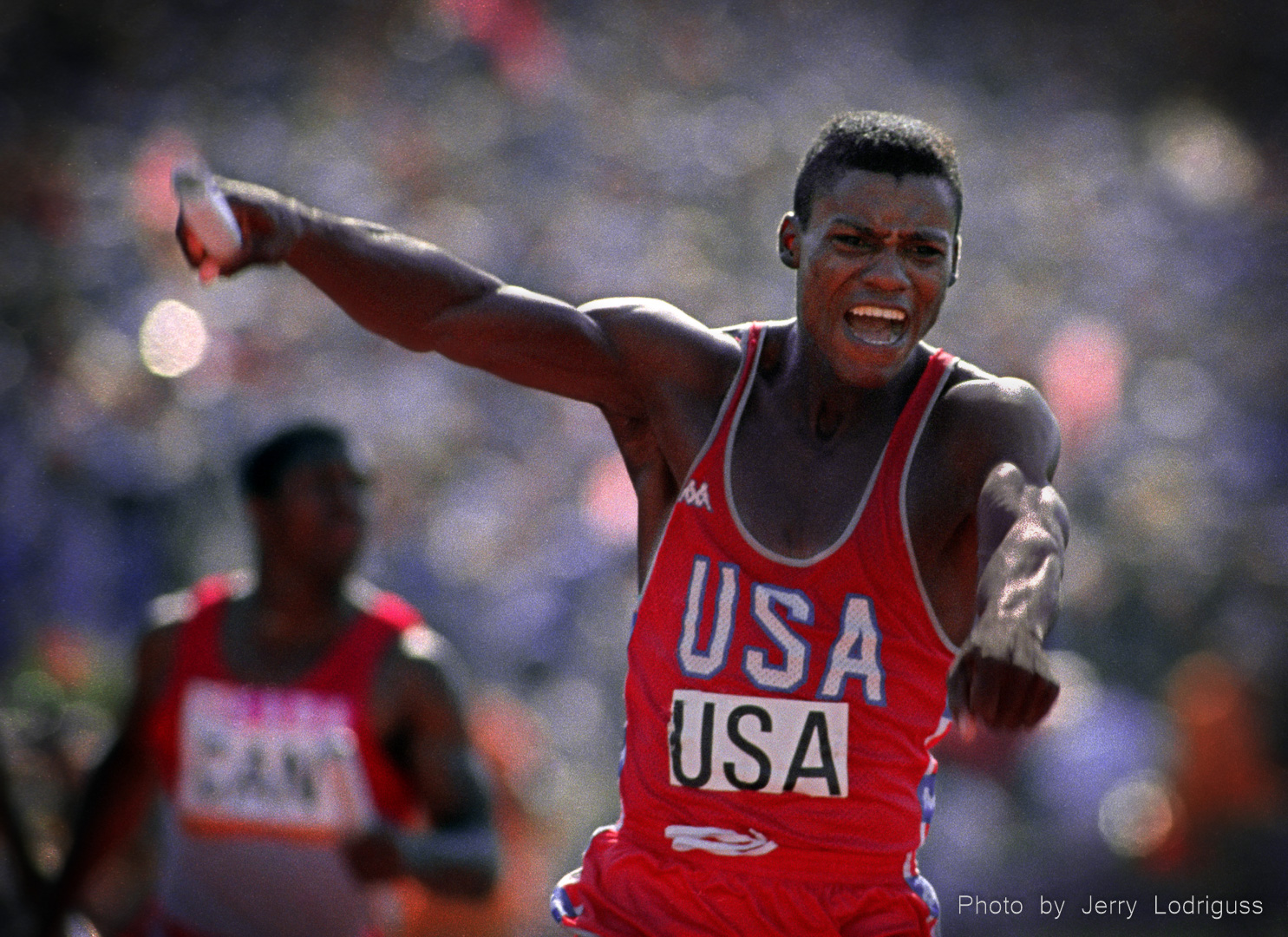 Carl Lewis reacts in joy after seeing a world record time posted on the scoreboard as he crosses the finish line in the men's 400 meter relay. Lewis won his 4th gold medal of the games when he anchored the relay team to a 37.83 clocking.
