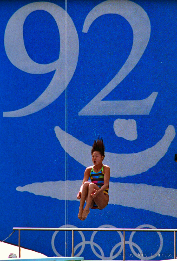 In front of a giant '92 Olympics sign, Gao Min of China spins during a dive making her hair stand on end. She won a gold medal in the finals of the women's 3 meter springboard competition during the 1992 Barcelona Olympics. Nicknamed the "Diving Queen", Gao is one of the most dominant divers in the history of the sport. Undefeated in world competition on the 3-meter springboard between 1986 and 1992 (including Olympic Games gold medals both in 1988 in Seoul and 1992 in Barcelona). She is tied with the legendary Greg Louganis in winning the greatest number of international awards on one board.